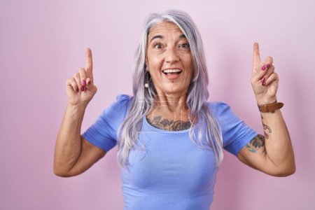 Foto de Mujer de mediana edad con tatuajes de pie sobre fondo rosa sonriendo sorprendido y sorprendido y apuntando hacia arriba con los dedos y los brazos levantados. - Imagen libre de derechos