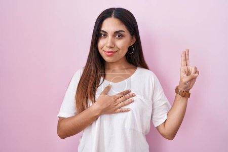 Téléchargez les photos : Jeune femme arabe debout sur fond rose souriant jurer avec la main sur la poitrine et les doigts vers le haut, faisant une promesse de fidélité serment - en image libre de droit
