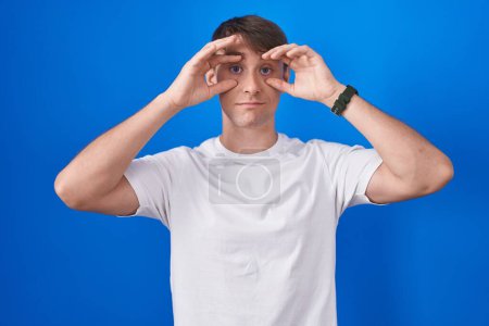 Photo for Caucasian blond man standing over blue background trying to open eyes with fingers, sleepy and tired for morning fatigue - Royalty Free Image