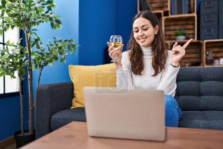 Photo for Young hispanic woman doing video call drinking white wine smiling happy pointing with hand and finger to the side - Royalty Free Image