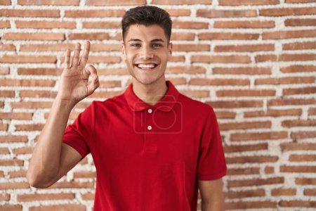 Photo for Young hispanic man standing over bricks wall smiling positive doing ok sign with hand and fingers. successful expression. - Royalty Free Image