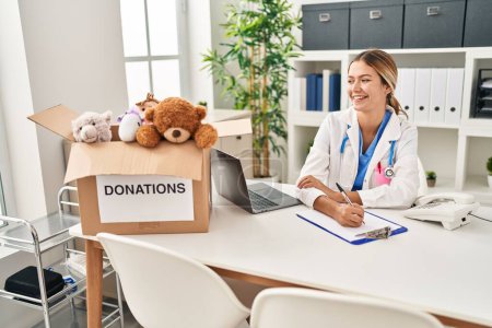 Photo for Young hispanic woman wearing doctor uniform writing report for donations toys at clinic - Royalty Free Image