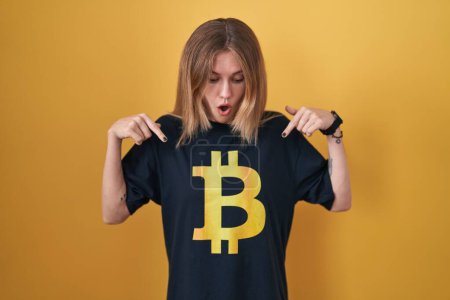 Téléchargez les photos : Blonde femme caucasienne portant Bitcoin t-shirt pointant vers le bas avec les doigts montrant la publicité, visage surpris et bouche ouverte - en image libre de droit
