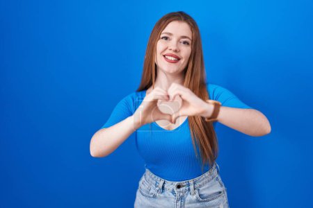 Photo for Redhead woman standing over blue background smiling in love doing heart symbol shape with hands. romantic concept. - Royalty Free Image