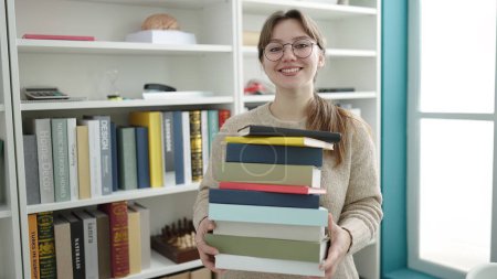 Téléchargez les photos : Jeune étudiante blonde souriante confiante tenant des livres à la bibliothèque de l'université - en image libre de droit