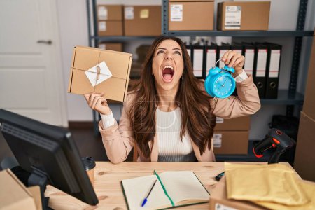 Photo for Young brunette woman working at small business commerce holding backpack angry and mad screaming frustrated and furious, shouting with anger looking up. - Royalty Free Image