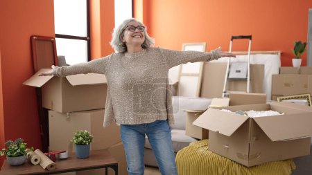 Téléchargez les photos : Femme d'âge moyen avec les cheveux gris souriant confiant debout avec les bras ouverts à la nouvelle maison - en image libre de droit
