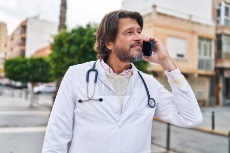 Téléchargez les photos : Moyen âge homme médecin souriant confiant parler sur smartphone à la rue - en image libre de droit