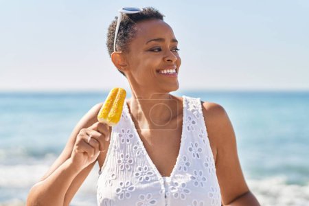 Téléchargez les photos : Femme afro-américaine souriante manger de la glace en toute confiance au bord de la mer - en image libre de droit