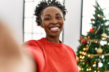 Foto de Mujer afroamericana hacer selfie por cámara de pie por árbol de Navidad en casa - Imagen libre de derechos