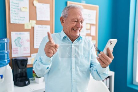 Téléchargez les photos : Homme âgé avec les cheveux gris travaillant au bureau faire appel vidéo avec smartphone souriant heureux pointant avec la main et le doigt sur le côté - en image libre de droit