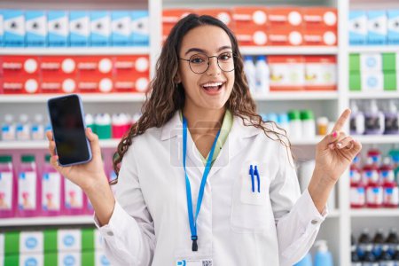 Photo for Young hispanic woman working at pharmacy drugstore showing smartphone screen angry and mad screaming frustrated and furious, shouting with anger looking up. - Royalty Free Image