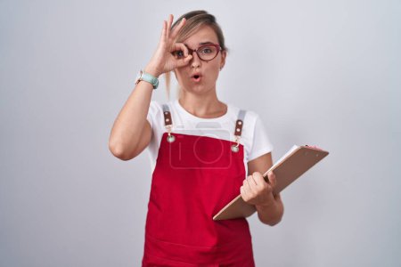 Photo for Young blonde woman wearing waiter uniform holding clipboard doing ok gesture shocked with surprised face, eye looking through fingers. unbelieving expression. - Royalty Free Image