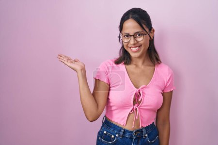 Téléchargez les photos : Jeune femme hispanique debout sur fond rose portant des lunettes souriant gai présentation et pointant avec la paume de la main regardant la caméra. - en image libre de droit