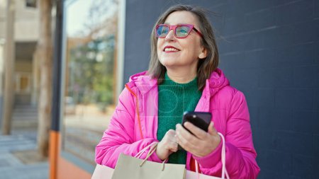 Foto de Mujer hispana madura con el pelo gris sonriendo yendo de compras usando smartphone en la calle - Imagen libre de derechos