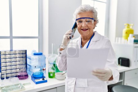 Foto de Mujer mayor de cabello gris con uniforme científico hablando en el teléfono inteligente en el laboratorio - Imagen libre de derechos
