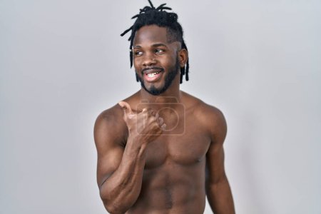 Foto de Hombre africano con rastas de pie sin camisa sobre fondo aislado sonriendo con la cara feliz mirando y señalando a un lado con el pulgar hacia arriba. - Imagen libre de derechos