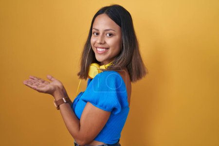 Foto de Mujer joven hispana de pie sobre fondo amarillo señalando a un lado con las manos abiertas palmeras mostrando espacio de copia, presentando anuncio sonriente emocionado feliz - Imagen libre de derechos