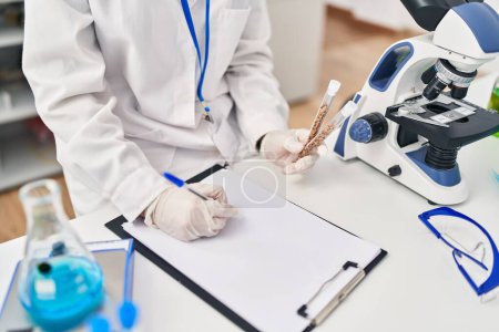 Téléchargez les photos : Young blonde woman wearing scientist uniform holding test tubes writing on checklist at laboratory - en image libre de droit