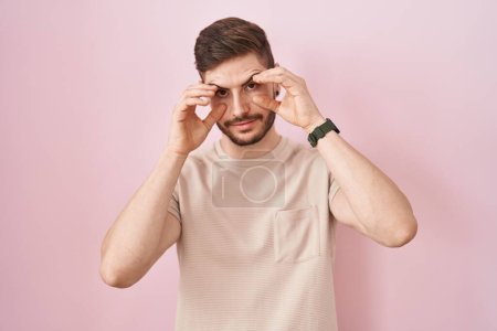 Foto de Hombre hispano con barba de pie sobre fondo rosa tratando de abrir los ojos con los dedos, somnoliento y cansado de fatiga matutina - Imagen libre de derechos
