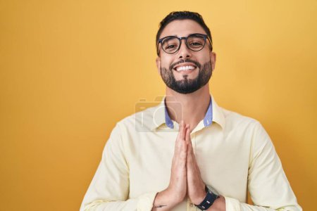 Téléchargez les photos : Jeune homme hispanique portant des vêtements de travail et des lunettes priant avec les mains ensemble demandant pardon souriant confiant. - en image libre de droit