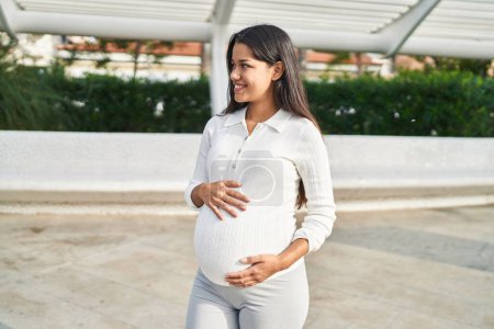 Téléchargez les photos : Jeune femme latine enceinte souriant confiant toucher le ventre à la rue - en image libre de droit