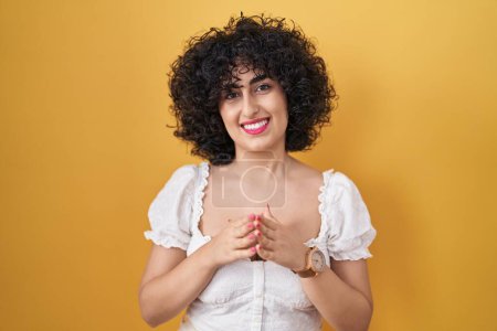 Téléchargez les photos : Jeune femme brune avec les cheveux bouclés debout sur fond jaune mains ensemble et doigts croisés souriant détendu et joyeux. succès et optimisme - en image libre de droit