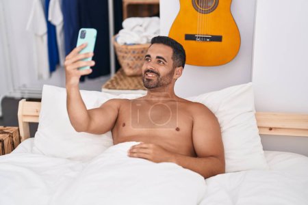 Téléchargez les photos : Young hispanic man make selfie by smartphone lying on bed at bedroom - en image libre de droit