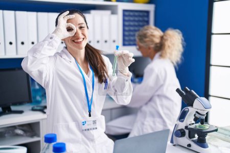Photo for Young hispanic woman working at scientist laboratory smiling happy doing ok sign with hand on eye looking through fingers - Royalty Free Image