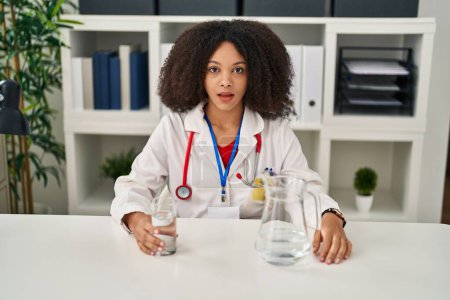 Foto de Joven africana americana médico mujer beber agua en la clínica asustado y sorprendido con la boca abierta para sorpresa, incredulidad cara - Imagen libre de derechos