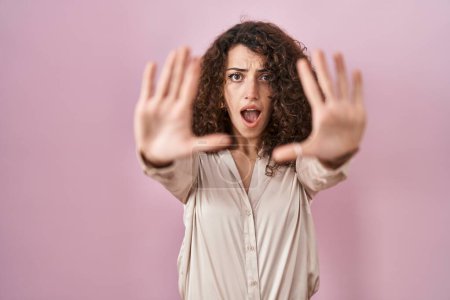 Photo for Hispanic woman with curly hair standing over pink background doing stop gesture with hands palms, angry and frustration expression - Royalty Free Image