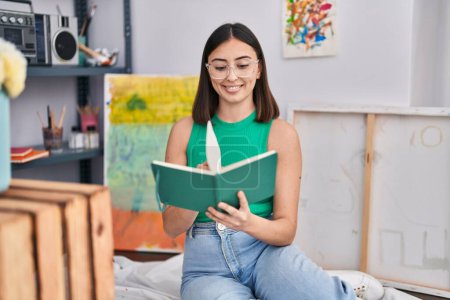 Photo for Young hispanic woman artist reading book at art studio - Royalty Free Image