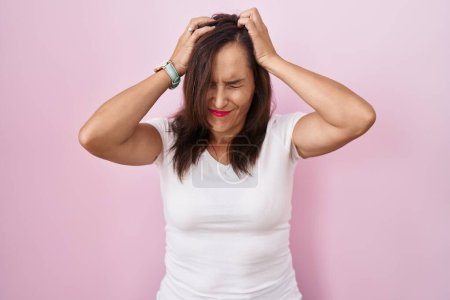 Téléchargez les photos : Femme brune d'âge moyen debout sur fond rose souffrant de maux de tête désespérés et stressés parce que la douleur et la migraine. mains sur la tête. - en image libre de droit