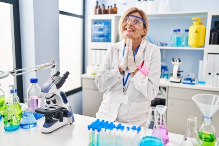 Photo for Middle age blonde woman working at scientist laboratory praying with hands together asking for forgiveness smiling confident. - Royalty Free Image