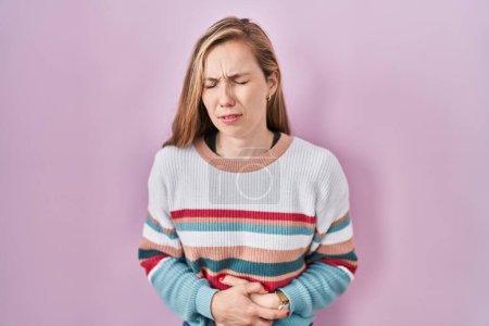 Foto de Young blonde woman standing over pink background with hand on stomach because indigestion, painful illness feeling unwell. ache concept. - Imagen libre de derechos