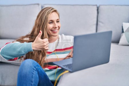 Téléchargez les photos : Jeune femme blonde utilisant un ordinateur portable à la maison souriant heureux et positif, pouce levé faire excellent et signe d'approbation - en image libre de droit