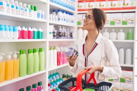 Téléchargez les photos : Jeune belle hispanique femme client souriant confiant tenant bouteille de shampooing et panier de marché à la pharmacie - en image libre de droit