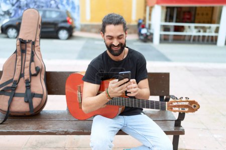 Téléchargez les photos : Jeune musicien hispanique tenant une guitare classique à l'aide d'un smartphone dans la rue - en image libre de droit