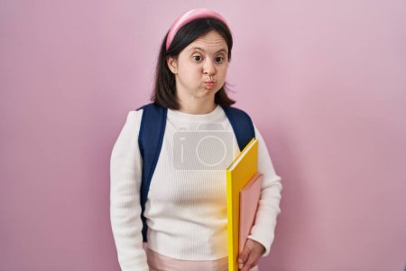 Téléchargez les photos : Femme avec le syndrome du duvet portant un sac à dos d'étudiant et tenant des livres gonflant les joues avec un visage drôle. bouche gonflée d'air, expression folle. - en image libre de droit