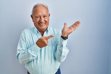 Foto de Hombre mayor con el pelo gris de pie sobre fondo azul asombrado y sonriendo a la cámara mientras se presenta con la mano y señalando con el dedo. - Imagen libre de derechos