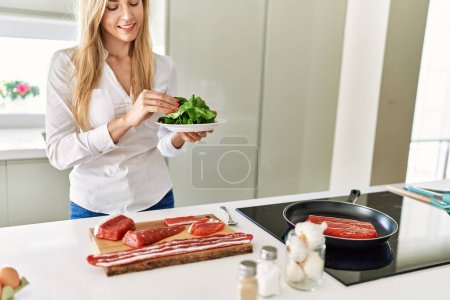Téléchargez les photos : Jeune femme blonde souriante confiante cuisine de bœuf à la cuisine - en image libre de droit
