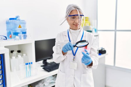 Téléchargez les photos : Femme aux cheveux gris d'âge moyen portant un uniforme de scientifique à la loupe au laboratoire - en image libre de droit