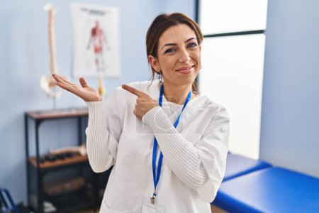 Téléchargez les photos : Jeune femme brune travaillant à la clinique de récupération de la douleur étonnée et souriante à la caméra tout en présentant avec la main et pointant du doigt. - en image libre de droit