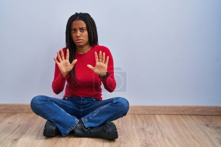 Foto de Joven afroamericano con trenzas sentadas en el suelo en casa alejando las palmas de las manos mostrando rechazo y negación con expresión de miedo y repugnante. parada y prohibido. - Imagen libre de derechos