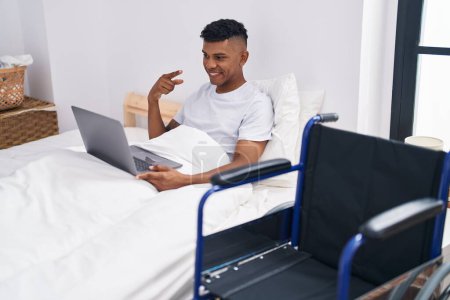 Photo for Young hispanic man lying on the bed, using wheelchair smiling happy pointing with hand and finger - Royalty Free Image