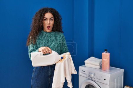 Foto de Mujer hispana joven sosteniendo ropa sucia y botella de detergente en la cara de choque, mirando escéptico y sarcástico, sorprendido con la boca abierta - Imagen libre de derechos