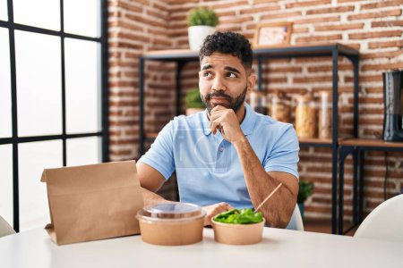 Photo for Hispanic man with beard eating delivery salad serious face thinking about question with hand on chin, thoughtful about confusing idea - Royalty Free Image