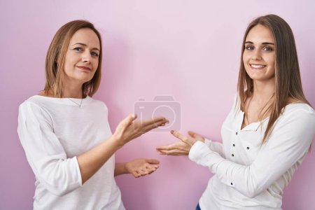 Photo for Middle age mother and young daughter standing over pink background inviting to enter smiling natural with open hand - Royalty Free Image