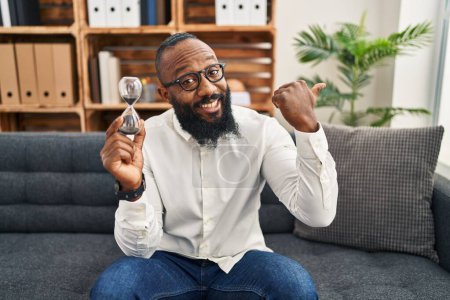 Foto de Hombre afroamericano sosteniendo reloj de arena en la oficina apuntando pulgar hacia el lado sonriendo feliz con la boca abierta - Imagen libre de derechos