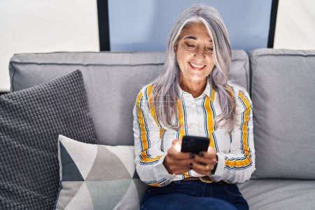 Téléchargez les photos : Femme aux cheveux gris du moyen âge utilisant un smartphone assis sur un canapé à la maison - en image libre de droit